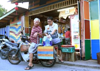 Anak Tan Cheng Hock,72, membantu mengangkat barang untuk diletakkan di atas motorsikal ayahnya di kedai runcit milik mereka di Kampung Nelayan Warna-Warni Seberang Ramai, Kuala Perlis, kelmarin. – UTUSAN/IZLIZAN OTHMAN