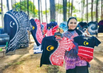 SEORANG  pekerja House Of Tie Dye, Shasuhaida Abdullah menunjukkan antara baju T ‘ikat dan celup’ yang dijual di kawasan riadah dan rekreasi Pantai Sura, Dungun, Terengganu, semalam. – UTUSAN/NIK NUR IZZATUL HAZWANI NIK ADNAN