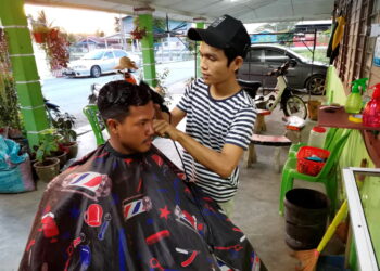 PEMUDA orang kurang upaya (OKU) Farid Aidil Mat Din menggunting rambut pelanggannya di rumahnya di Kampung Teluk Bakong, Lambor Kanan dekat Parit ketika ditemui hari ini. -UTUSAN/AIN SAFRE BIDIN