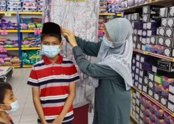 RODZIAH Mohd. Razali (kanan) mengenakan songkok di kepala kedua orang murid anak yatim ketika berbelanja pakaian baharu hari raya di sebuah pasar raya bandar Jerantut, Jerantut, Pahang. - UTUSAN/HARIS FADILAH AHMAD