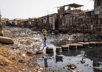 SEORANG wanita berjalan atas timbunan tayar ketika melintasi terusan air yang dicemari sampah di kawasan setinggan di Freetown, Sierra Leone. - AFP