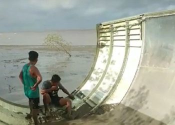 DUA penduduk duduk di atas logam yang didakwa serpihan pesawat di Pantai Teluk Ranggau, Kalimantan Tengah. -KOMPAS.COM